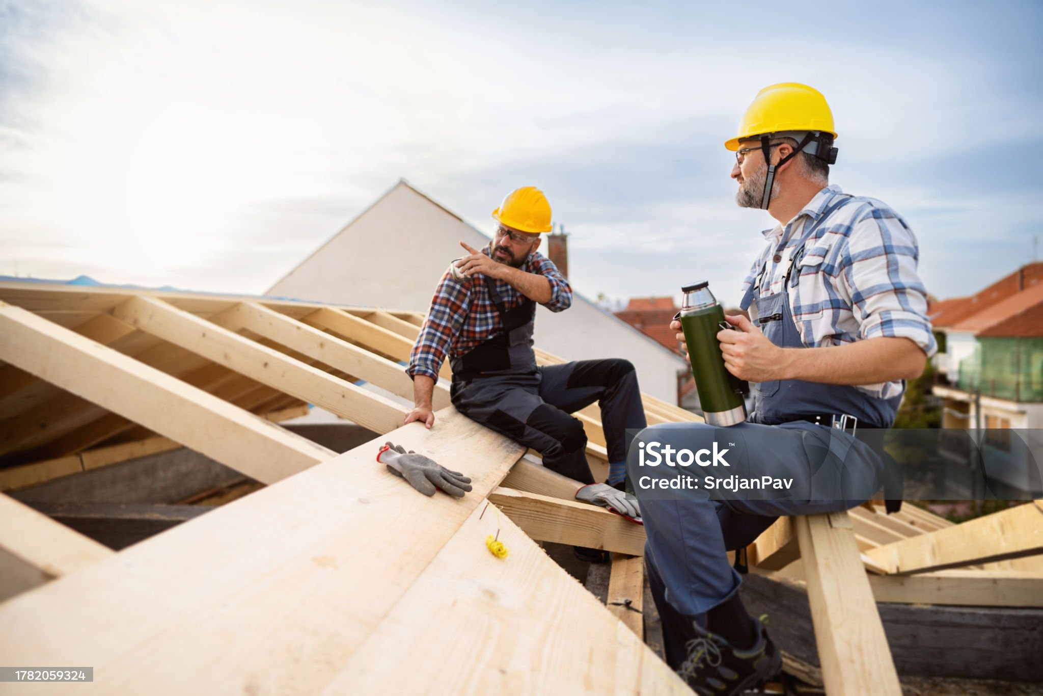 Two mid-adult male Caucasian roofers, taking an break from work while sitting on a roof beam, and drinking coffee or water